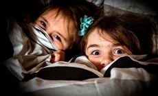 girls hiding under the bedcovers - one scared, one smiling